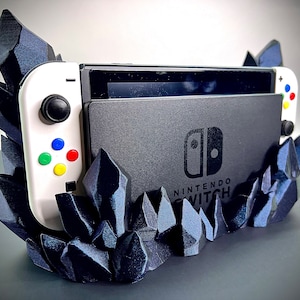 May include: A black Nintendo Switch console and white controllers are displayed in a dark blue, crystal-shaped holder. The holder has a geometric design and the Nintendo Switch logo is visible.