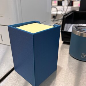 May include: A blue desk organizer with a yellow sticky note dispenser. The organizer is made of a textured material and has a square shape.