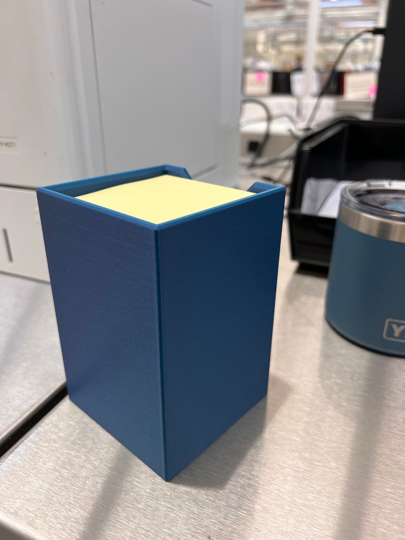 May include: A blue desk organizer with a yellow sticky note dispenser. The organizer is made of a textured material and has a square shape.
