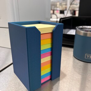 May include: A blue plastic holder with a stack of colorful sticky notes. The notes are yellow, orange, pink, light blue, green, and light yellow.