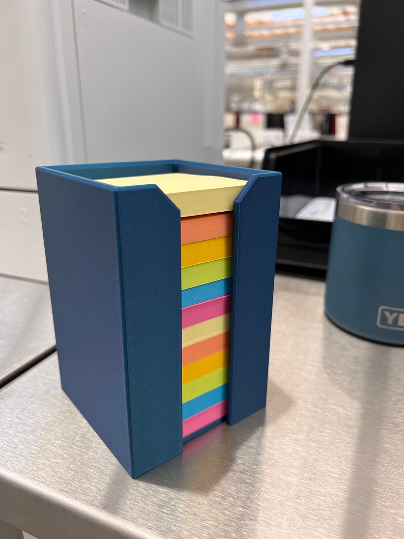 May include: A blue plastic holder with a stack of colorful sticky notes. The notes are yellow, orange, pink, light blue, green, and light yellow.
