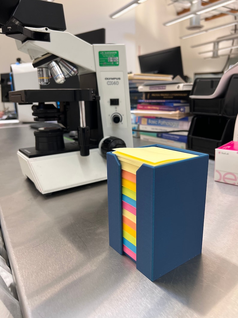 May include: A blue plastic holder with a stack of colorful sticky notes. The holder is on a silver surface next to a white microscope.