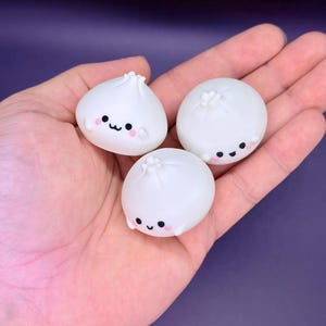 Puede incluir: Tres pequeñas figuras blancas con forma de dumpling, con caras sonrientes y mejillas rosadas. Los dumplings tienen una superficie texturizada y están dispuestos en una mano sobre un fondo púrpura. Las figuras tienen un diseño lindo y caprichoso.