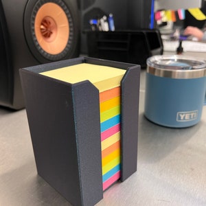 May include: A black, rectangular, 3D printed holder for sticky notes. The holder has a slot for the notes to be dispensed from. The notes are multi-colored and are stacked in the holder. The holder is sitting on a silver surface.