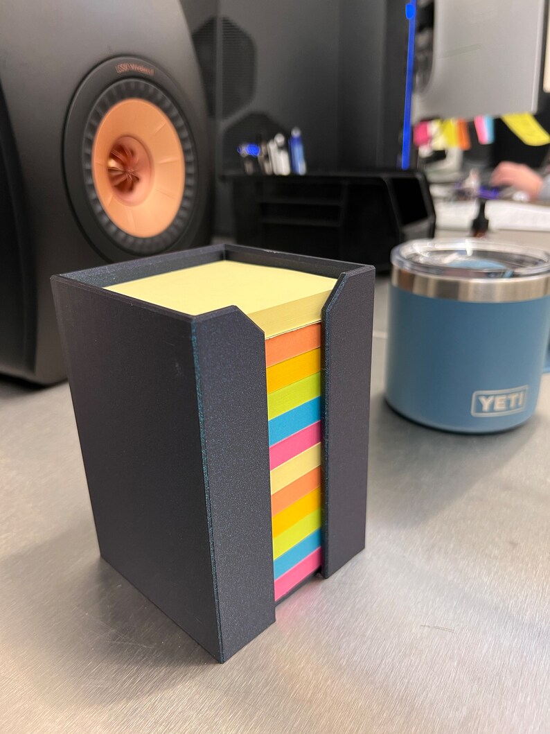 May include: A black, rectangular, 3D printed holder for sticky notes. The holder has a slot for the notes to be dispensed from. The notes are multi-colored and are stacked in the holder. The holder is sitting on a silver surface.
