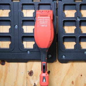 May include: A red and black power tool with the text "FUEL" is mounted on a black storage system. The tool has a long handle and a red head. The storage system is attached to a wooden surface.