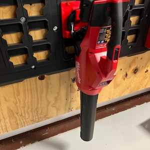 May include: A red and black Milwaukee leaf blower mounted on a black wall organizer. The blower is attached to a red bracket. The wall organizer is mounted on a wooden surface.