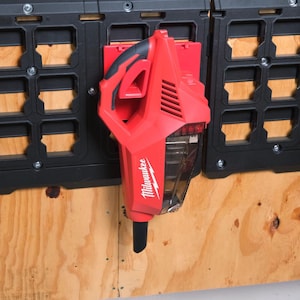 May include: A red Milwaukee cordless vacuum cleaner mounted on a black storage panel. The vacuum has a clear dust collection chamber and a black nozzle. The storage panel is attached to a wooden surface.