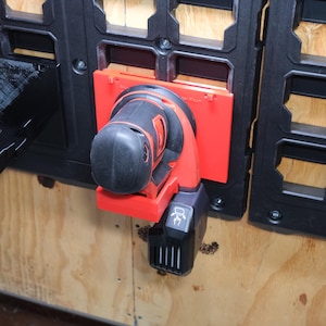 May include: A red and black power sander mounted on a red bracket, attached to a black storage system. The sander is positioned against a light-colored wooden surface. The storage system has a grid-like design.