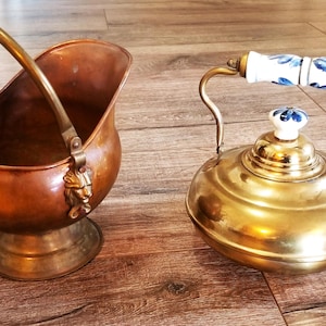 May include: A copper coal scuttle and a brass kettle. The scuttle has a handle and a lion's head detail. The kettle is gold-coloured with a spout and a lid with a floral knob. The kettle handle is white with blue floral details.