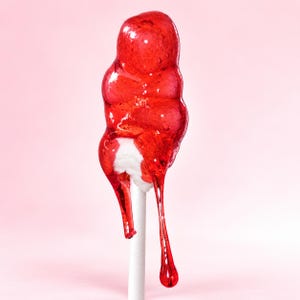 Könnte beinhalten: Ein durchscheinender roter Lutscher mit glänzendem, geschmolzenem Aussehen, der einen weißen Kern bedeckt. Das Bonbon hat eine abgerundete, unregelmäßige Form mit einem dicken, zähflüssigen Tropfen, der an einem dünnen, geraden weißen Stiel hängt.