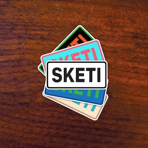 May include: Three colorful stickers with the word "SKEITI" printed in white on a black background. The stickers are layered on top of each other, with the top sticker being green, the middle sticker being pink, and the bottom sticker being blue.