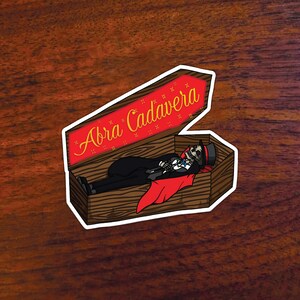 May include: A sticker of a skeleton in a coffin with the text "Abra Cadavara" on the lid. The coffin is brown wood with a red lining. The skeleton is wearing a black suit, a red cape, and a top hat.