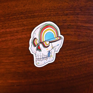 May include: A white skull sticker with a rainbow design emerging from the top of the skull. The sticker has a blue opening and colorful paint splatters. The sticker is on a dark brown wooden surface.