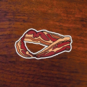 May include: A sticker featuring a cartoon illustration of bacon strips. The bacon is depicted with red and tan colors, with a white border. The sticker is placed on a dark brown wooden surface.
