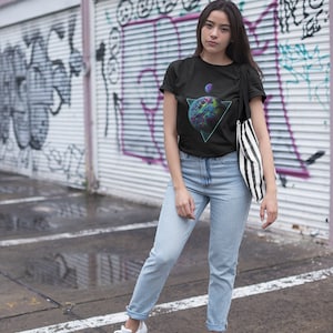 May include: A woman wearing a black t-shirt with a graphic of a planet in a triangle. The graphic is in shades of blue, green, and purple. She is also wearing light blue jeans and white sneakers. She is standing in front of a graffiti-covered wall.