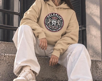 Puede incluir: Una sudadera con capucha de color beige que presenta un gráfico circular en el pecho. El diseño incluye el texto RELA4LIFE y FREEDOM IS A STATE OF MIND, rodeando un escudo central con una corona, dos leones y el número 7 en un escudo.