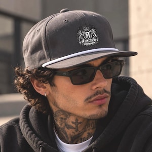 Puede incluir: Una gorra de béisbol gris carbón con un detalle de cuerda blanca en la visera y un logotipo central de escudo con el texto RELA4LIFE, una corona y dos leones. La gorra presenta una corona estructurada y una visera plana de color sólido.
