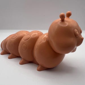 May include: A peach-colored plastic caterpillar toy with a segmented body and a face with closed eyes.