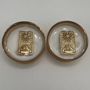 May include: Two gold-colored round medallions with a clear glass top. Each medallion contains a gold-colored rectangular plaque with a sunburst design and a small diamond in the center.