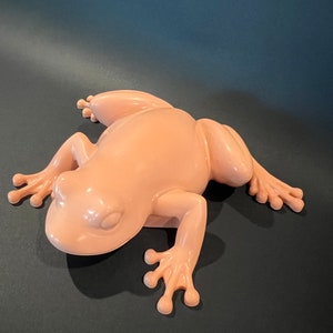 May include: A light pink ceramic frog figurine with a smooth, glossy finish. The frog is posed in a sitting position with its legs outstretched.