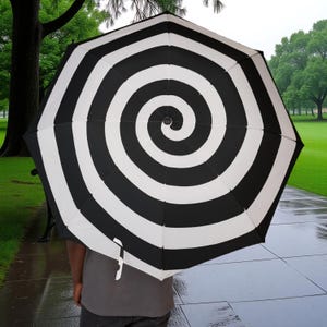 Puede incluir: Paraguas con diseño de espiral en blanco y negro. El diseño del paraguas presenta rayas curvas negras y blancas alternas que crean un efecto de espiral. El mango es visible.