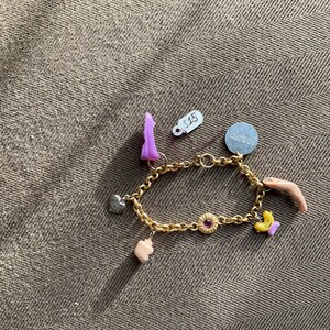 May include: Gold-toned charm bracelet with various charms, including a heart, a purple shape, a flower, and a small hand. The bracelet is on a textured brown surface.