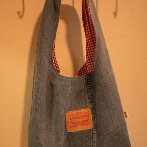 Op de afbeelding: Een hobo-schoudertas gemaakt van blauwe spijkerstof. De binnenkant is gevoerd met een rood-wit gingham ruitpatroon. Een beige leren merklabel van Levi Strauss & Co. is op de voorzijde van de spijkerstof tas genaaid.