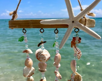 May include: A wooden seashell wind chime with a starfish hanging from the top. The chime features multiple strands of beads and seashells in shades of pink, white, and blue.