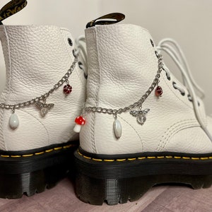 Puede incluir: Botas blancas de cuero con cadenas y abalorios plateados, incluyendo setas, una mariposa y una abeja. Las botas tienen suelas y cordones negros.