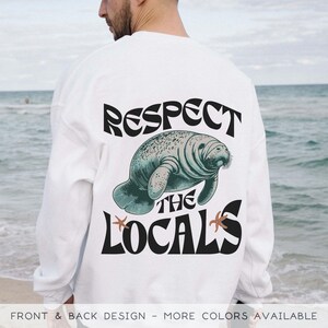 May include: White long-sleeve sweatshirt with a graphic of a manatee and the text "Respect the Locals" in black retro font.