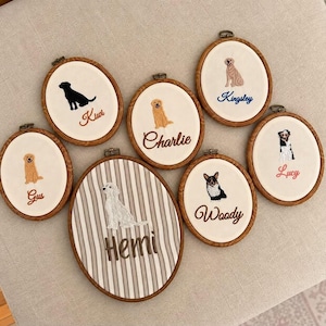 Könnte beinhalten: Sieben ovale Holzstickrahmen mit Stoff, auf dem Hundeporträts und Namen gestickt sind. Die Namen lauten Kiwi, Charlie, Kingsley, Gus, Hemi, Woody und Lucy. Jeder Rahmen zeigt eine individuelle Hundeillustration und den jeweiligen Namen.