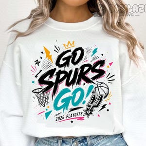 May include: A white crewneck sweatshirt featuring a graphic print with the black text GO SPURS GO, a teal GO, a basketball, a basketball hoop, and the text 2026 PLAYOFFS. The design includes yellow, teal, and pink accents with various geometric shapes.