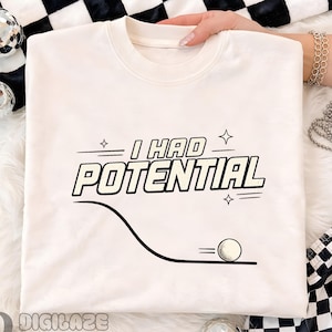 Puede incluir: Sudadera color crema con el texto "I HAD POTENTIAL" en negrita y en color negro. Debajo del texto hay un gráfico de una pelota de golf rodando por una línea curva. El diseño incluye pequeños acentos de estrellas.