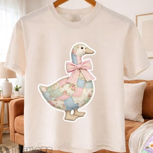 May include: Cream-colored t-shirt featuring a patchwork goose design. The goose has a pink bow and is made of various patterned fabrics, including floral and gingham prints. The shirt is hanging on a wooden hanger.