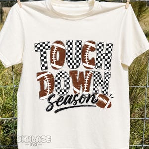 May include: A cream-colored short-sleeve t-shirt featuring the text TOUCH DOWN SEASON in a mix of black and white checkered patterns and brown football-themed graphics. The design includes football illustrations and the text DIGILAZE SVG.