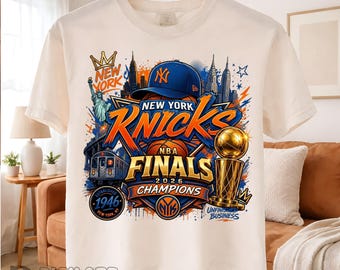 May include: A white t-shirt featuring a graphic print of New York Knicks NBA Finals 2026 Champions. The design includes a blue baseball cap, a basketball, the Statue of Liberty, a subway train, a trophy, and the text New York, Knicks, 1946.