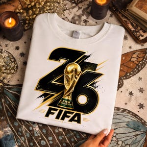 Może przedstawiać: Biała bluza z nadrukiem graficznym na Mistrzostwa Świata FIFA. Projekt zawiera numer "23" w kolorze czarnym i złotym, trofeum Mistrzostw Świata FIFA i słowo "FIFA".