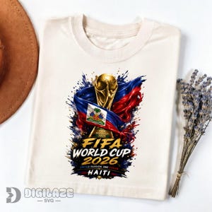 May include: A cream-colored t-shirt featuring a graphic of the FIFA World Cup 2026 trophy wrapped in a Haitian flag. The design includes the text FIFA WORLD CUP 2026, UNITED PASSION ONE DREAM, and HAITI, set against a vibrant blue and red paint splatter.