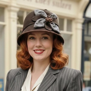 May include: A brown cloche hat with a gray flower accent. The hat has a rounded crown and a small brim. The woman in the image is wearing a gray pinstripe blazer and has red hair.