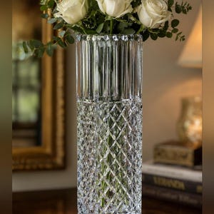 May include: A tall, clear glass vase with a square base and a multifaceted design. The lower half features a diamond-cut pattern, while the upper half has vertical fluted grooves. A bouquet of white roses and green eucalyptus leaves fills the vase.