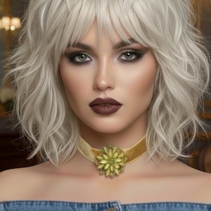 May include: A close-up of a person wearing a gold velvet choker necklace with a large, ornate flower pendant. The flower pendant is made of green and yellow gemstones. The person has light skin, gray hair, and dark eye makeup.