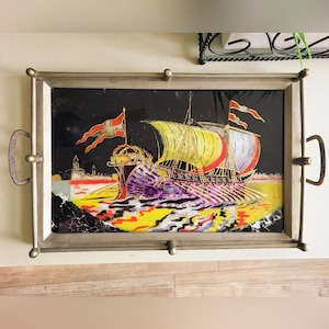 May include: Rectangular serving tray with a decorative ship scene. The artwork features a ship with yellow and red sails, red flags, and a black background. The tray has a silver metal frame with handles and decorative ball feet.