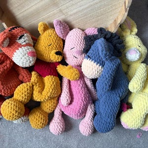 May include: A collection of five plush crochet stuffed animals featuring a tiger with orange and black stripes, a yellow bear with a red shirt, a pink piglet, a blue donkey with a black mane, and a yellow rabbit with pink inner ears.
