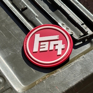 May include: A round, red and white patch featuring the Toyota logo in white. The patch is circular with a white border and is set against a green metal surface.