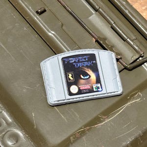 May include: A gray Nintendo 64 video game cartridge for "Perfect Dark" rests on a green metal surface. The cartridge features the game's title and a close-up of an eye.