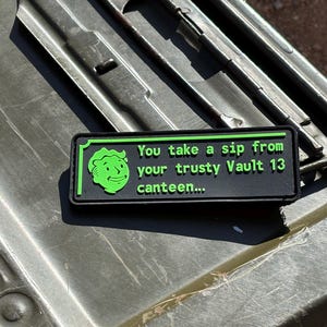 May include: A black and green patch with the text "You take a sip from your trusty Vault 13 canteen..." and a green cartoon face. The patch is rectangular and has a green border. The patch is on a metal surface.