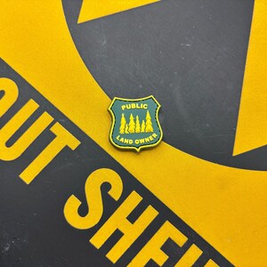 May include: A shield-shaped patch with a dark green background and a yellow border. The text PUBLIC LAND OWNER is printed in yellow capital letters. A graphic of five yellow pine trees is centered between the two lines of text.