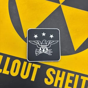 May include: A square black patch with a white border featuring a white graphic of an eagle with spread wings, a shield on its chest, and arrows in its talons, positioned below three white stars arranged in a horizontal arc across the top.