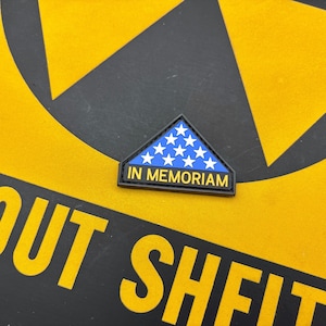 May include: A triangular patch featuring a blue field with white stars arranged in a folded flag pattern, with the text IN MEMORIAM in yellow capital letters on a black background. The patch has a black border and a flat, molded surface design.